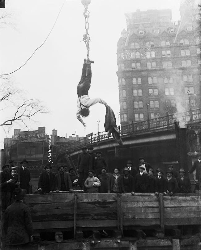 Spectators watch as escape artist Harry Houdini frees himself from a straightjacket while he hangs from a hook above a subway. Getty Images.