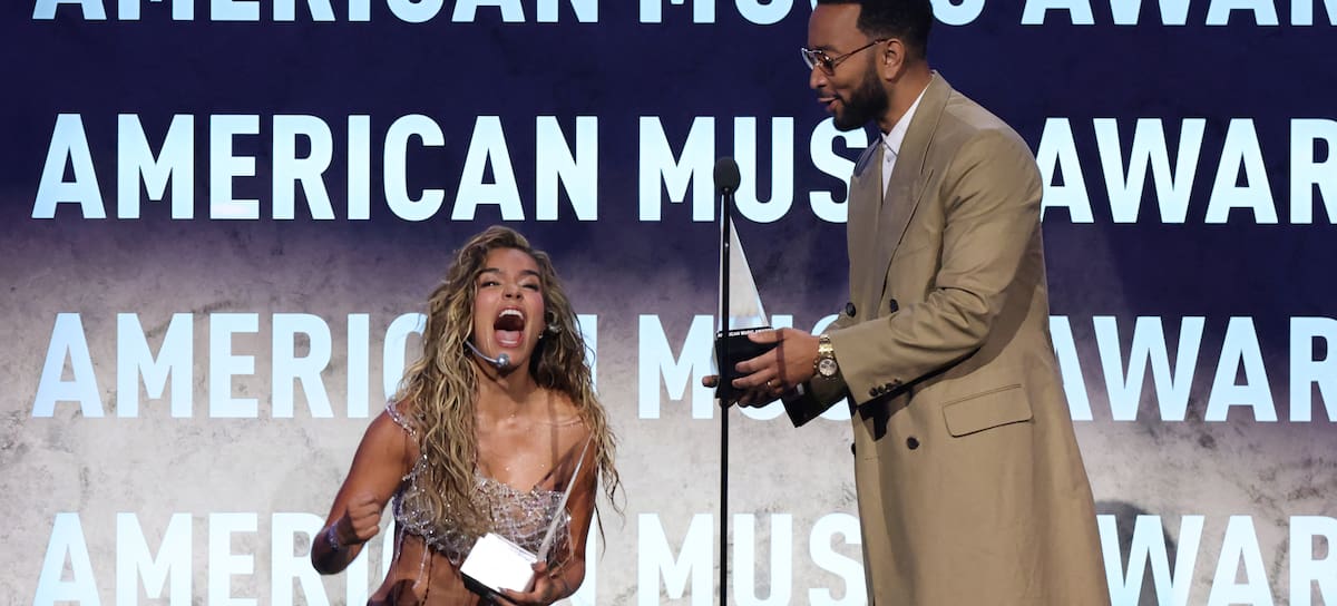 Colombian singer Karol G reacts as US singer-songwriter John Legend (R) presents her with the International Artist Award of Excellence and Best Latin Album for "Tropicoqueta" during the American Music Awards at the MGM Grand Garden Arena in Las Vegas, Nevada on May 25, 2026. (Photo by VALERIE MACON / AFP via Getty Images)