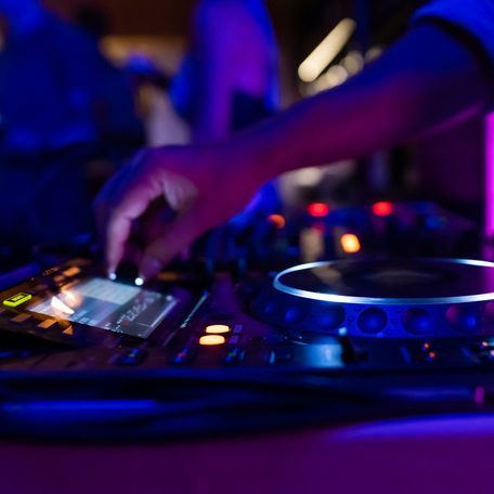 A DJ on a party mixing, controlling the volume and making music on stage with DJ equipment during the night in a colourful setting.