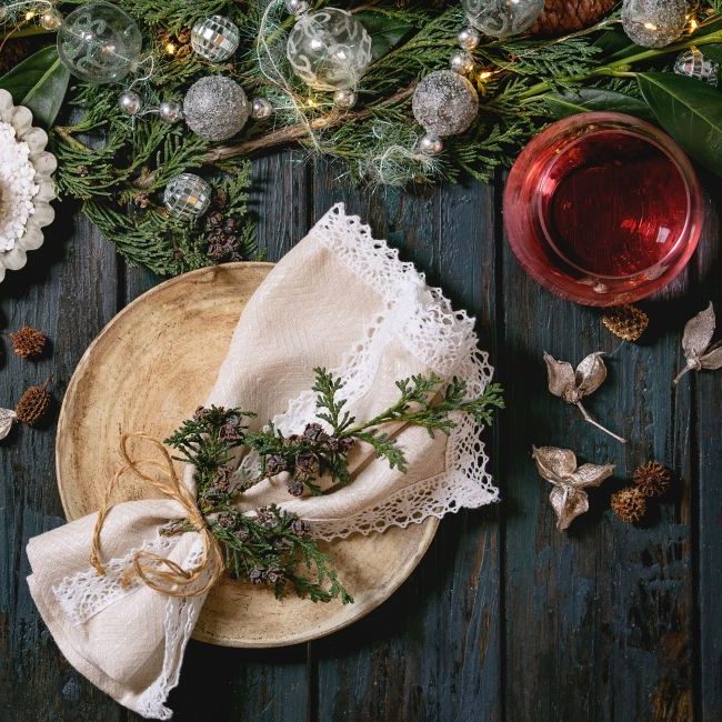 Mesa navideña o de Año Nuevo con platos de cerámica vacíos, copas de vino, servilletas, corona navideña de tuya, guirnalda luminosa y velas encendidas sobre una mesa de madera oscura. Decoración navideña.
