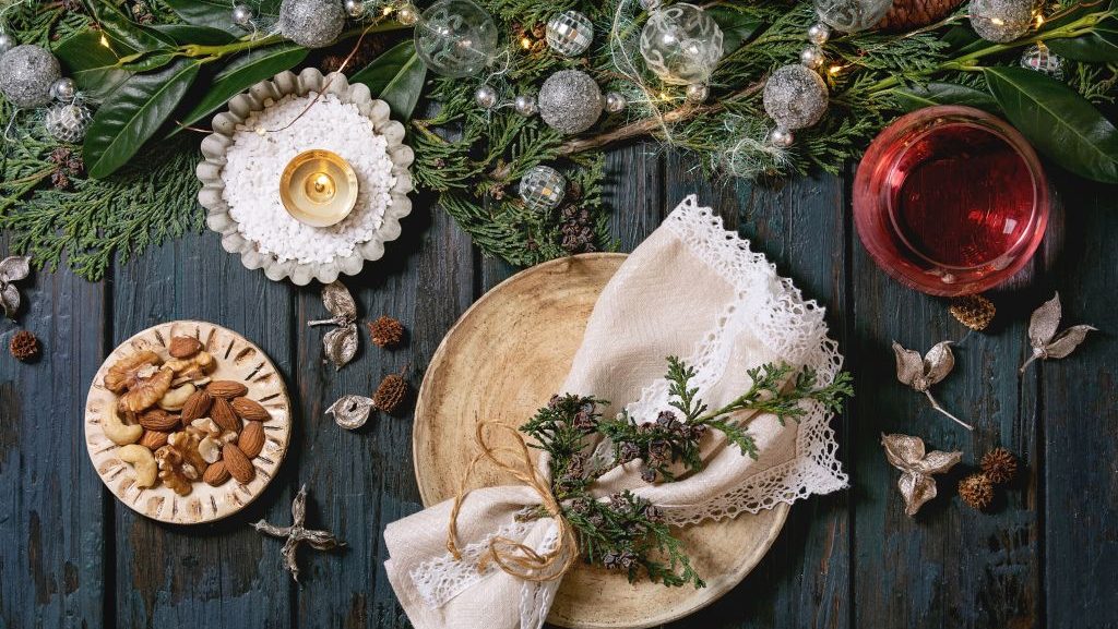 Mesa navideña o de Año Nuevo con platos de cerámica vacíos, copas de vino, servilletas, corona navideña de tuya, guirnalda luminosa y velas encendidas sobre una mesa de madera oscura. Decoración navideña.